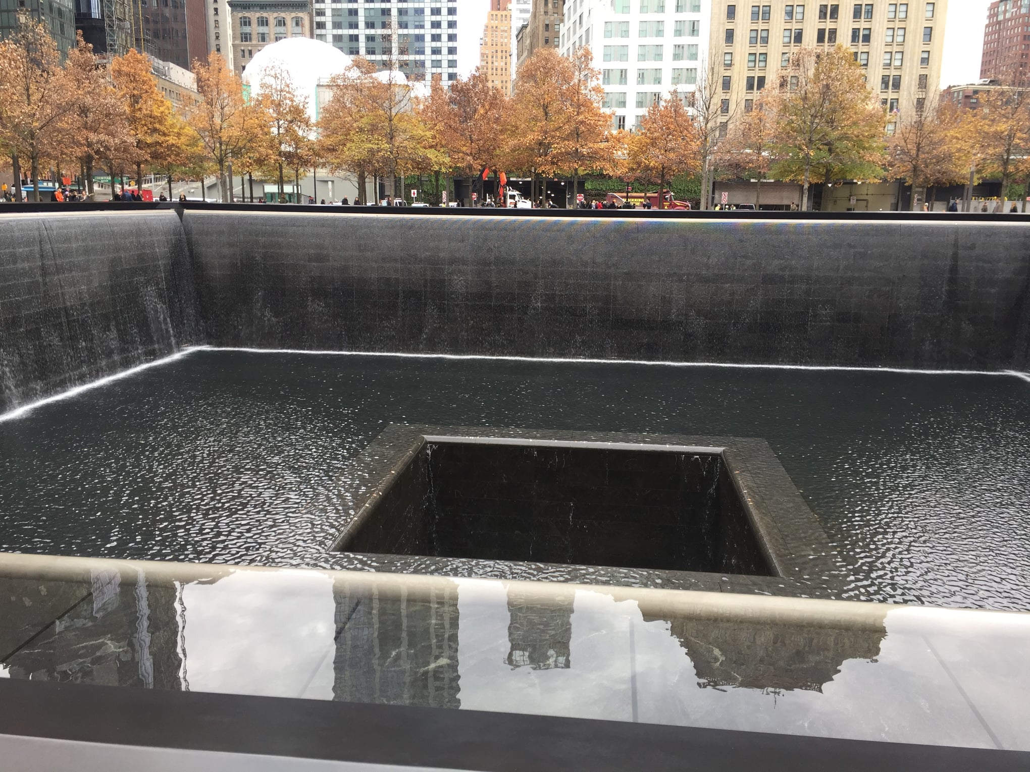 Water flows down four walls and disappears into a void at the center of a Memorial reflecting pool. Sunlight creates shadows on the pooled water.
