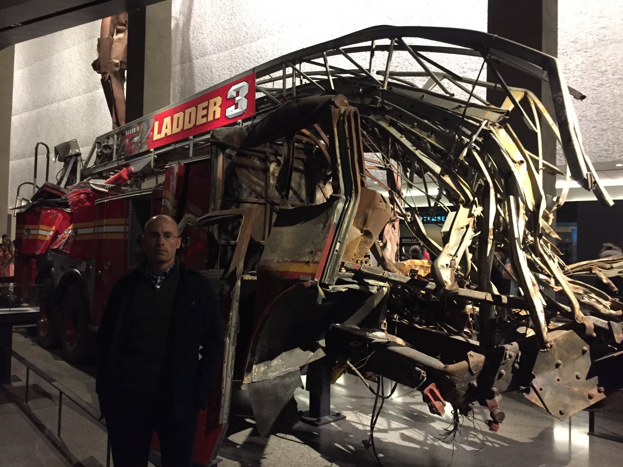 Tim Farmer (ARO) besides the heavily damaged firetruck of Ladder Company 3 sits in the Museum. This close-up view shows the bright red vehicle’s twisted ladder and broken compartment doors.